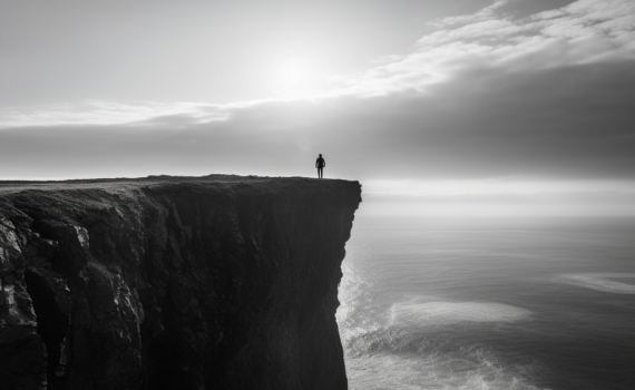 A grayscale image of a person standing at the edge of a cliff, looking out into the horizon. The scene depicts a poignant contrast between light and shadow, mirroring the extreme mood swings experienced by individuals with bipolar disorder and depression