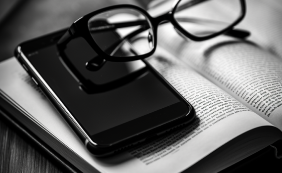 A striking black and white image featuring a pair of glasses resting on an open book. In the background, a smartphone lies, screen down, symbolizing the digital disconnect